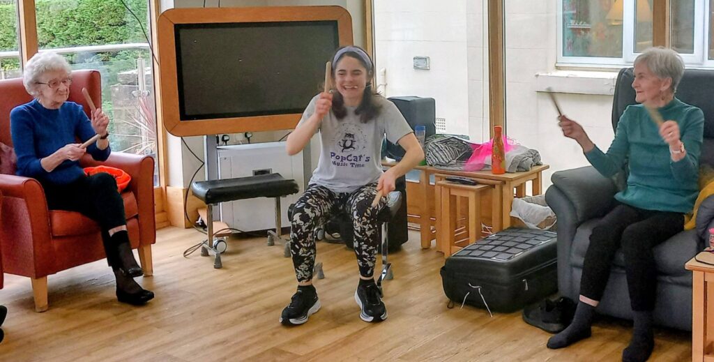 Three women sit in a bright room, smiling as they take part in a seated music activity, each holding either drumsticks or shakers.