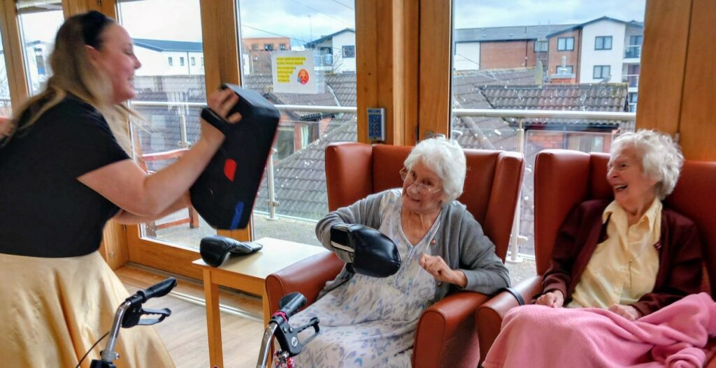 During an Olympic-themed session, a woman holds up boxing pads for an elderly woman wearing boxing gloves, whilst another elderly lady sits beside them, smiling.