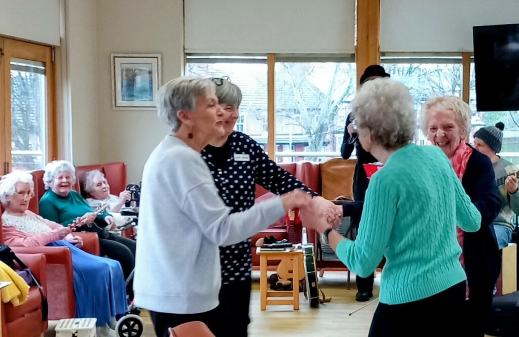 Several elderly women are dancing in time and smiling in a communal room, while others sit and watch from settees in the background, enjoying the lively atmosphere.