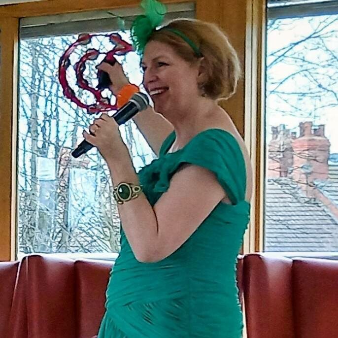 Sandra Currie, wearing a green dress, holds a microphone and a red decorative object during a cheerful visit, standing in front of a red sofa and large windows.