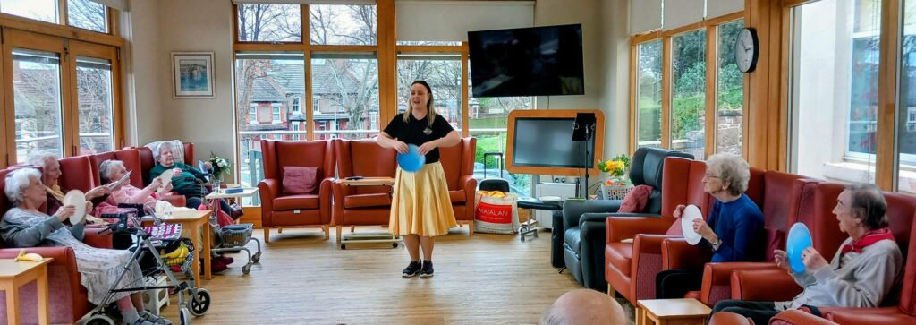 A woman stands smiling in the centre of a bright room during an Olympic-themed session as several elderly people sit in chairs around her, holding coloured paddles.