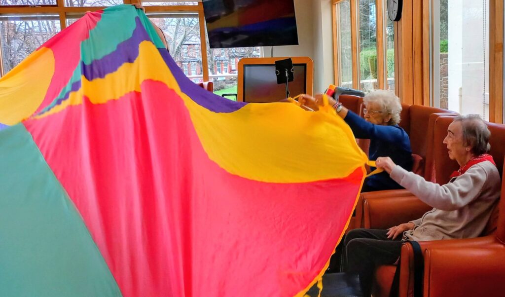 Two elderly people seated in armchairs hold up a large, colourful parachute indoors, enjoying an Olympic-themed group activity session.