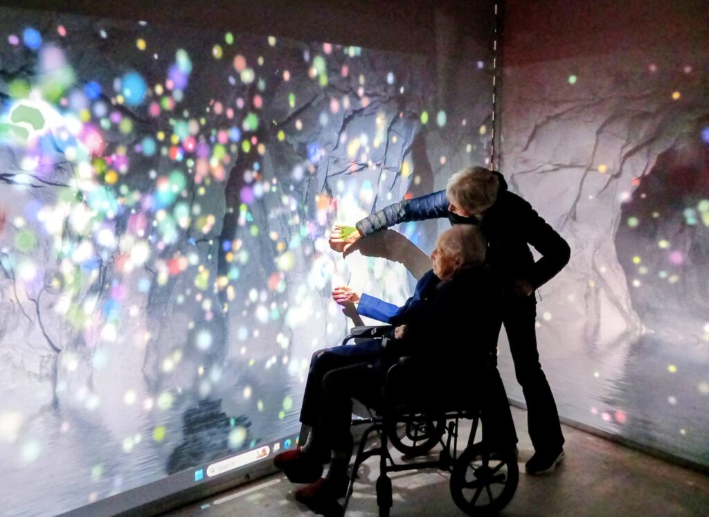 Two people, one using a wheelchair, engage with a colourful, projected digital display—journeying back in time amidst light spots and abstract patterns at the House of Memories.