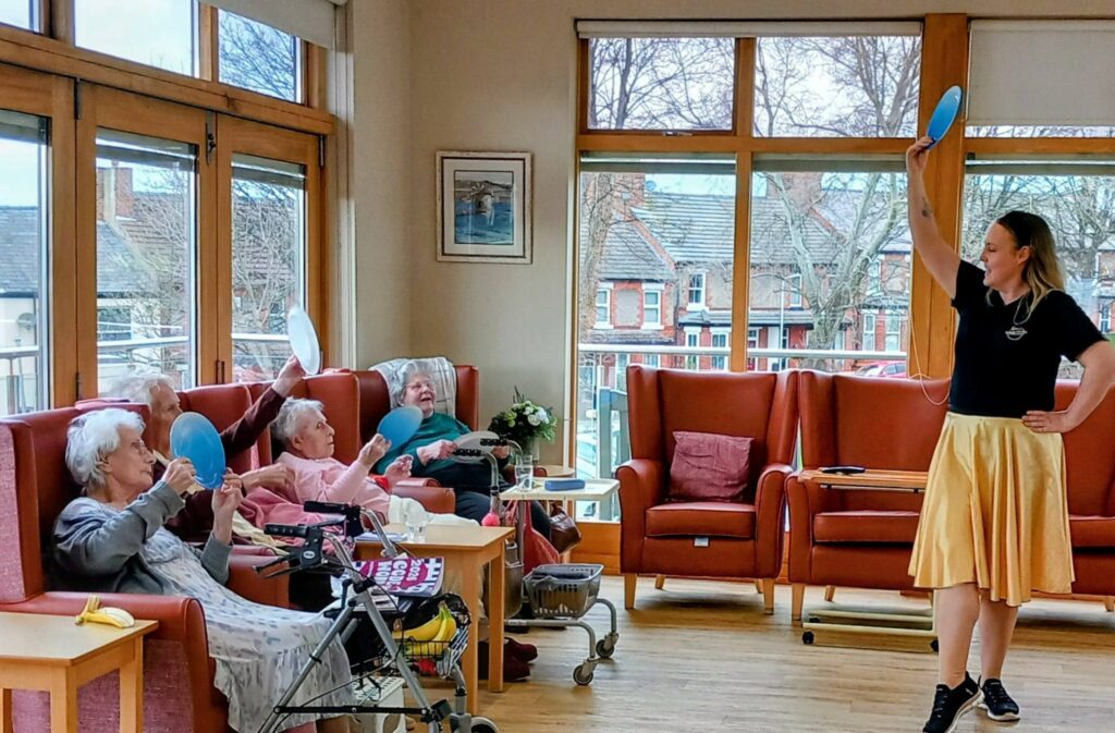 A group of elderly people seated in a care home take part in an Olympic-themed balloon activity session, led by a woman standing and holding a blue balloon.