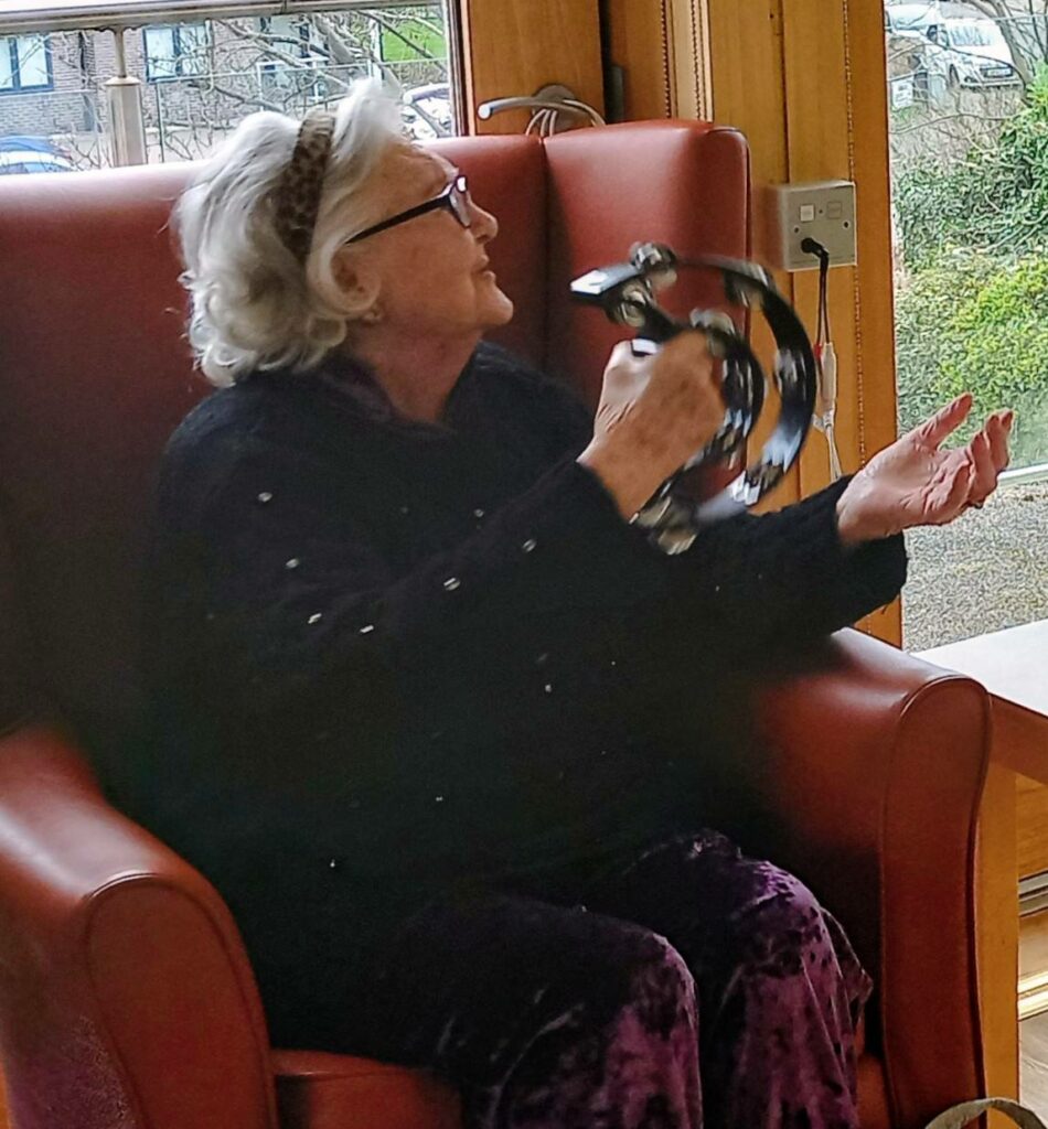 Sandra Currie, an older woman with grey hair and glasses, sits in a red armchair by a window, playing a black tambourine and looking to her left during a joyful visit.