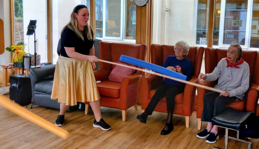 A woman in a gold skirt leads two elderly women in an Olympic-themed, seated music and movement session using foam sticks in a lounge area furnished with armchairs.