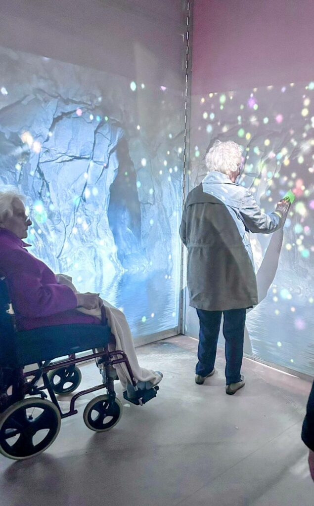 Two elderly women explore a light and projection display at the House of Memories; one is seated in a wheelchair whilst the other stands, touching the illuminated wall, Stepping Back in Time.