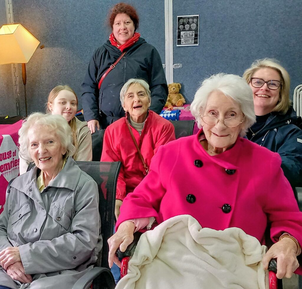 Six women of different ages, some sitting and some standing, pose and smile for the camera indoors, evoking nostalgia with a lamp and teddy bear in this House of Memories.