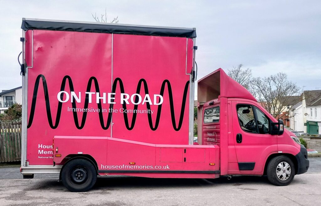 A bright pink House of Memories van with "ON THE ROAD Immersive in the Community" and houseofmemories.co.uk printed on the side, parked on a street, inviting you to journey back in time.