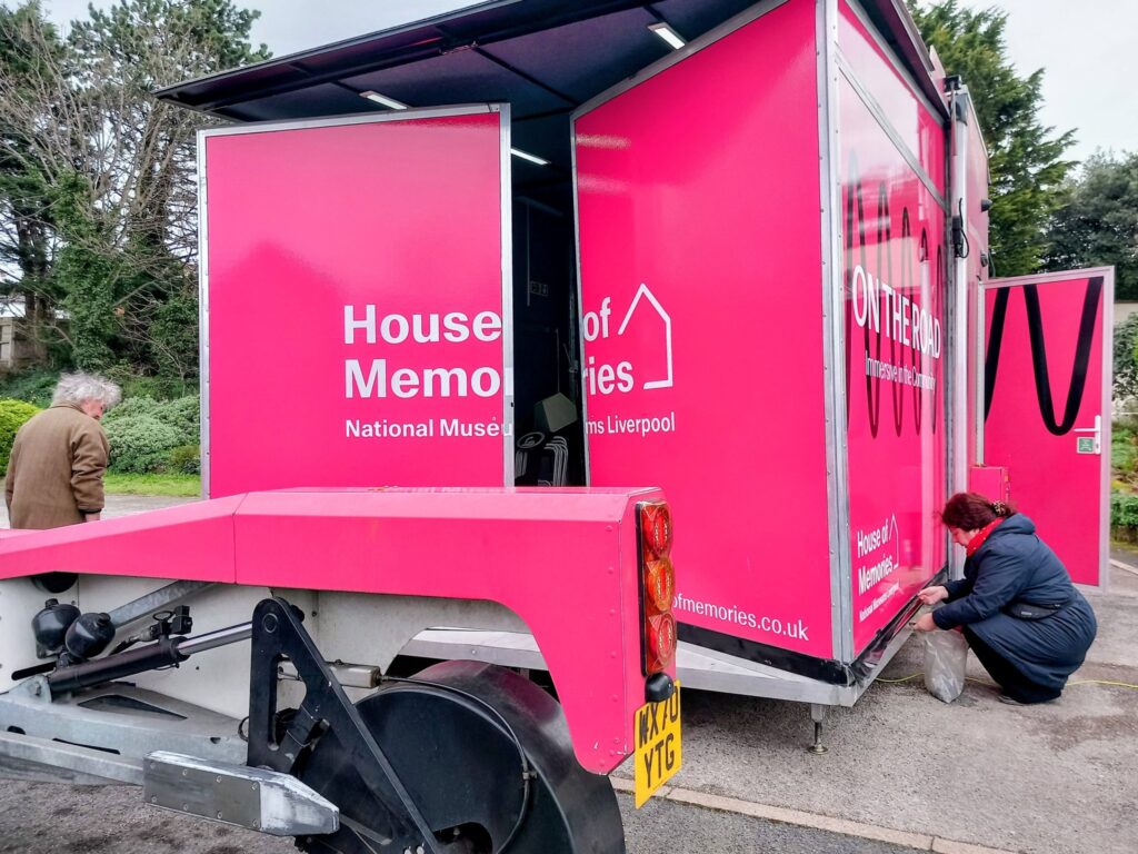 Two people step back in time next to a bright pink House of Memories mobile exhibition lorry from National Museums Liverpool, parked along a tree-lined street.