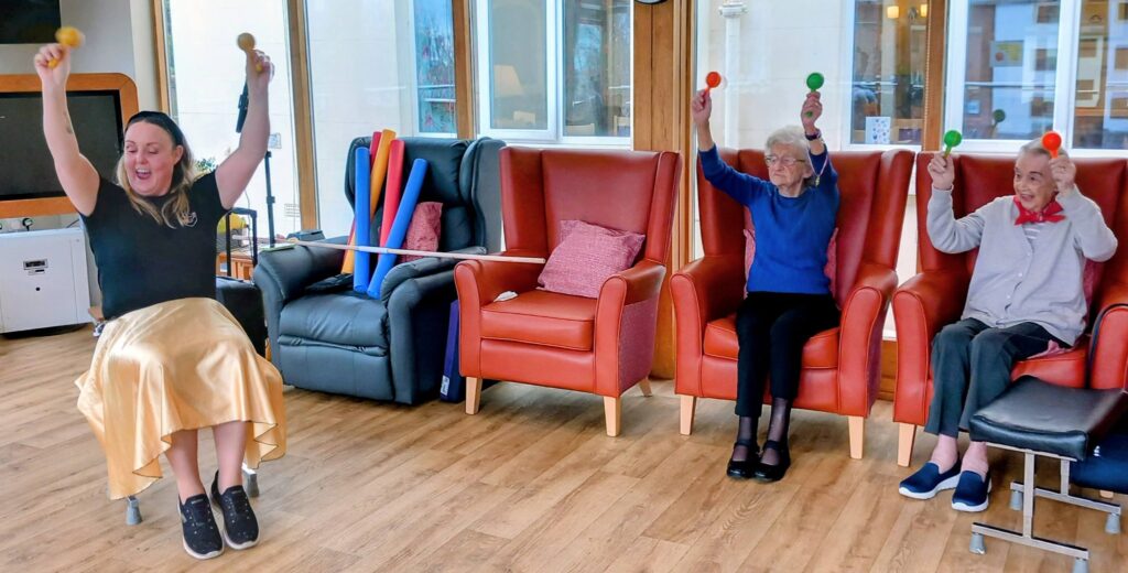 Three people are seated indoors on chairs, each holding up a colourful maraca during a music and movement session inspired by an Olympic-themed group activity.