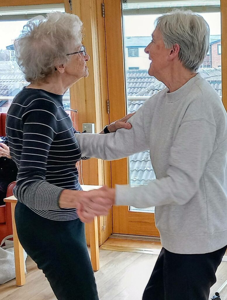 Two elderly women are holding hands and dancing together indoors, smiling at one another during a joyful visit.
