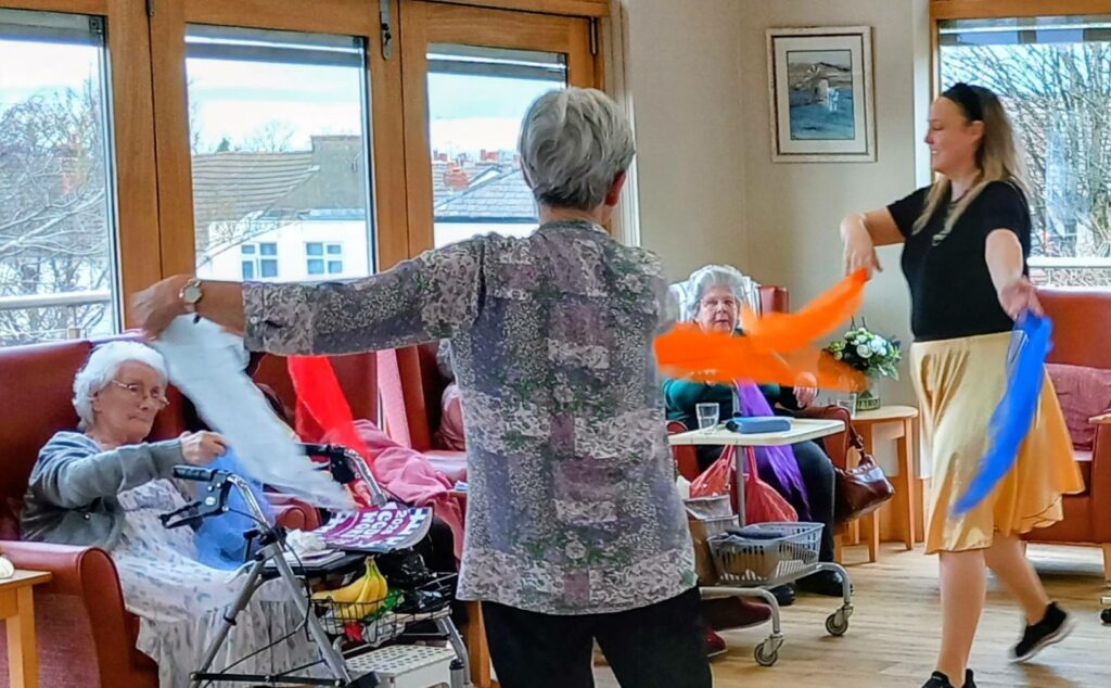 Two elderly women sit in a lounge whilst another woman and an instructor stand, holding colourful scarves and taking part in an Olympic-themed music and movement session.