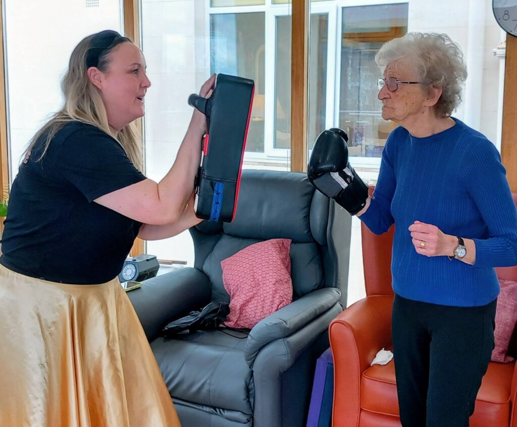 An elderly woman wearing boxing gloves practises punching a padded target held by another woman during an Olympic-themed session in a lounge setting.