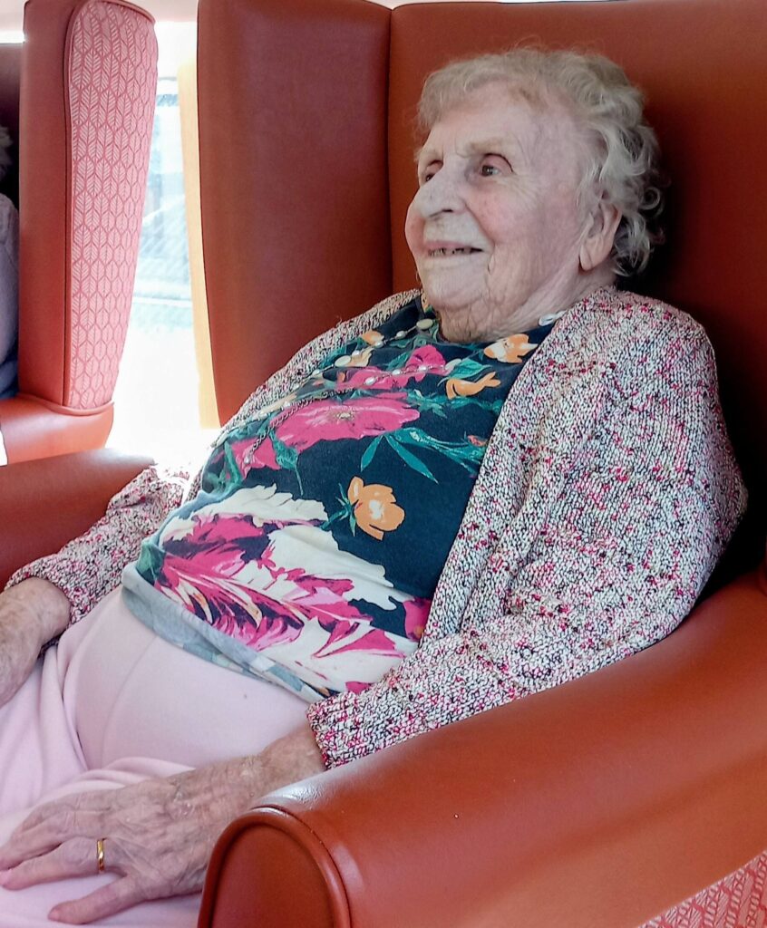 An elderly woman with short white hair sits in an orange armchair, dressed in a floral blouse, light pink trousers, and a patterned cardigan. Sandra Currie smiles warmly, clearly enjoying her visit as she looks slightly to her right.