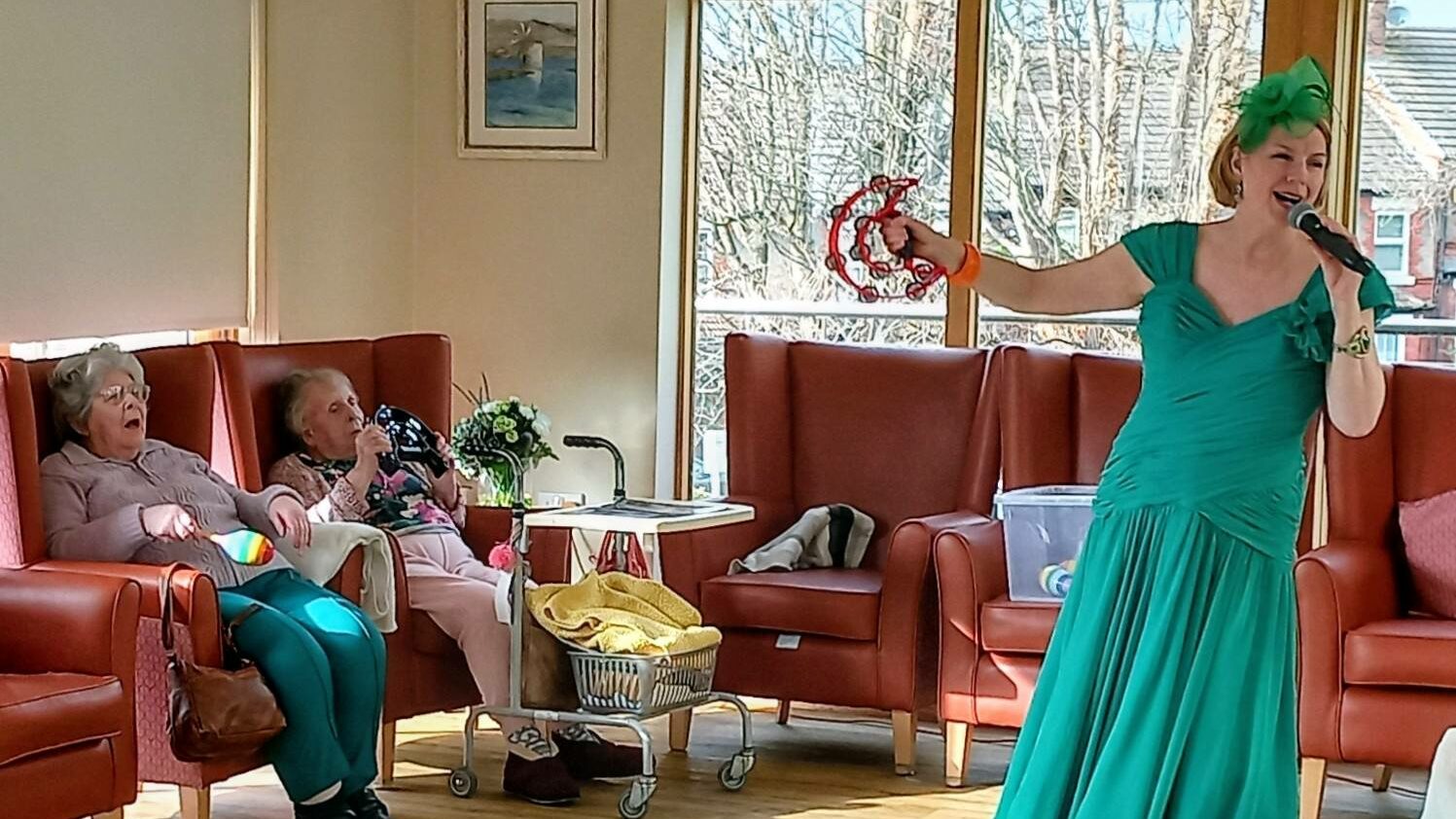 During a joyful visit, a woman in a green dress sings and plays the tambourine for two elderly women seated in armchairs in a brightly lit room.