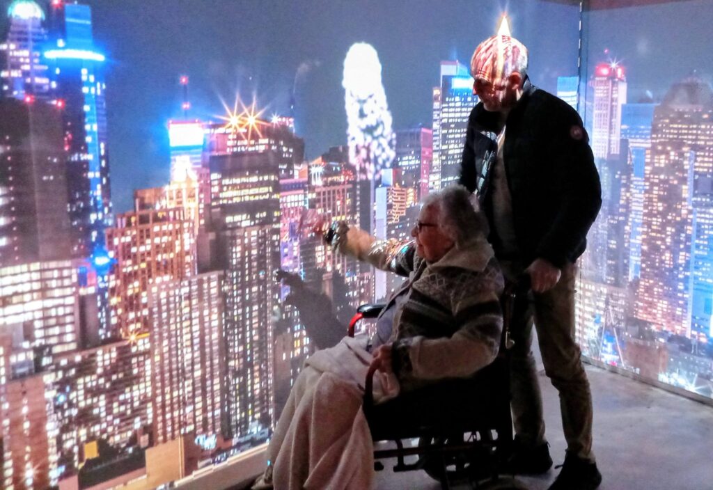 An older adult in a wheelchair gestures towards a cityscape projection, stepping back in time at the House of Memories, whilst another person stands beside them facing the illuminated city backdrop.