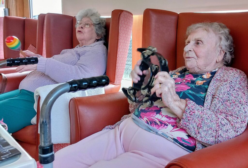 During a joyful visit, two elderly women sit in red armchairs, each holding a musical instrument—one with a maraca and the other with a tambourine. A mobility aid is visible in the foreground.