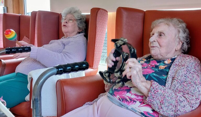 During a joyful visit, two elderly women sit in red armchairs, each holding a musical instrument—one with a maraca and the other with a tambourine. A mobility aid is visible in the foreground.