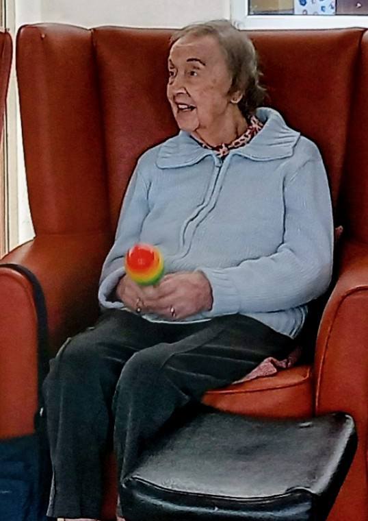 Elderly woman seated in a red armchair, holding a colourful maraca, wearing a light blue jumper and dark trousers during Sandra Currie’s joy-filled visit.