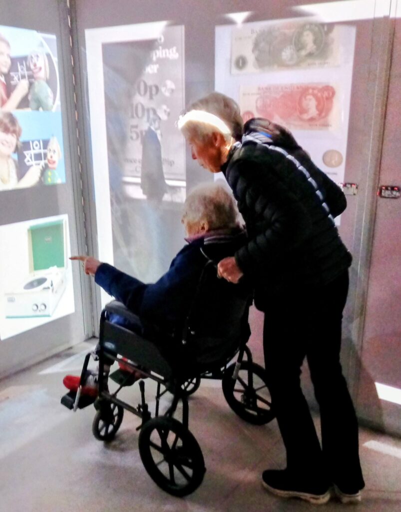 An elderly person in a wheelchair points at a display case, accompanied by another individual beside them in a museum, as they experience nostalgia and feel as though they're stepping back in time at the House of Memories.