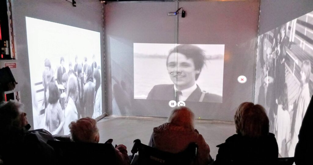 Four people sit in a dark room at the House of Memories, travelling back in time as black-and-white footage of crowds and a man in a suit is projected onto three walls.