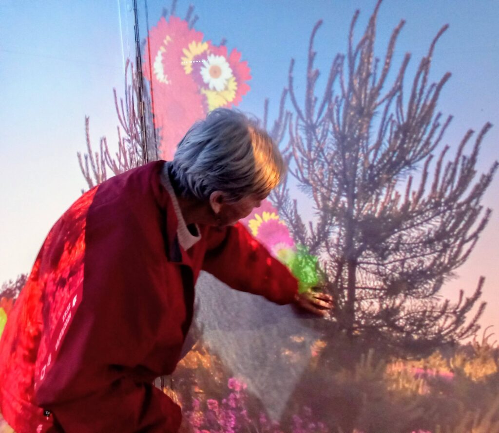 An older person in a red jacket touches a wall featuring a nature scene, stepping back in time at the House of Memories, surrounded by a tree and colourful flowers.