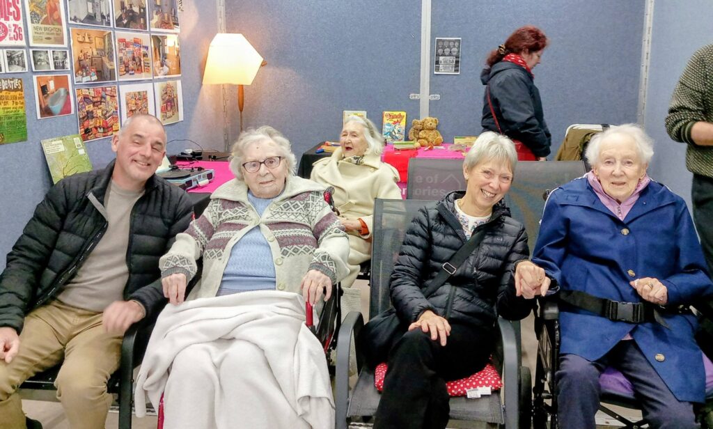 Four pensioners and a younger adult sit indoors, smiling as if reminiscing; posters and a lamp adorn the background. Another person stands behind them, turned away, creating a nostalgic House of Memories atmosphere.