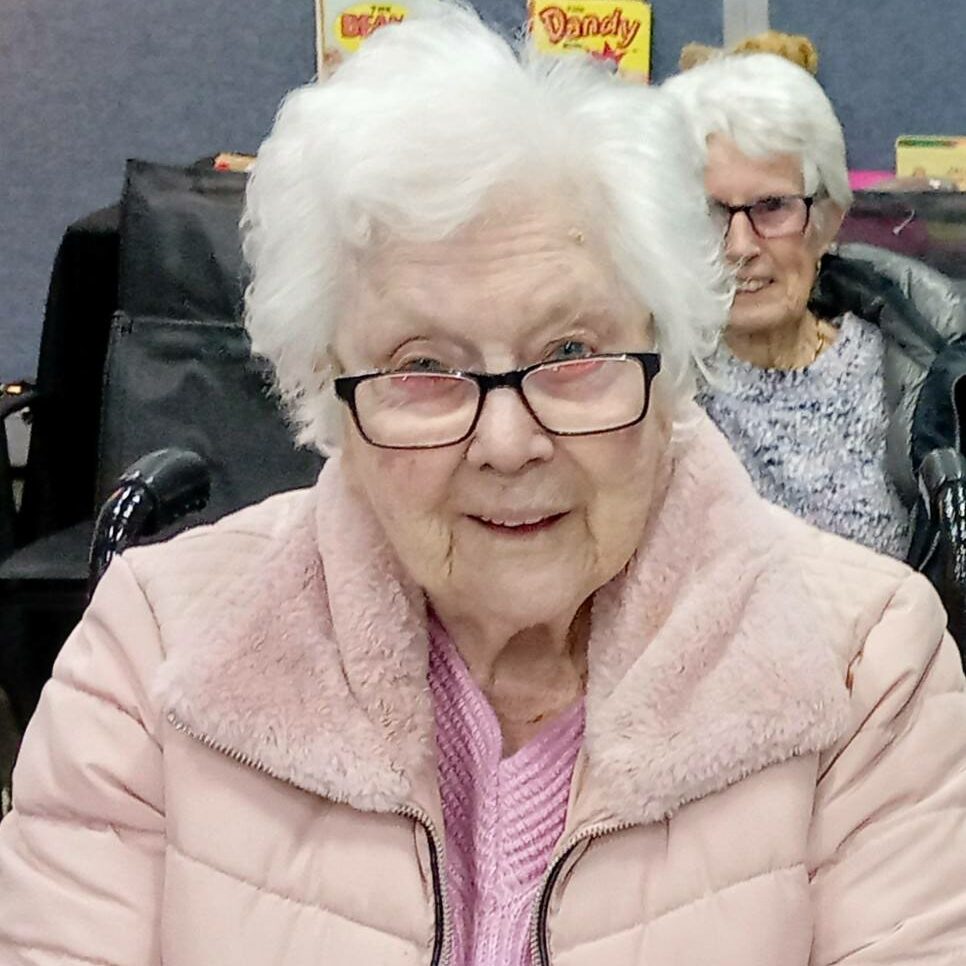 An elderly woman with white hair and glasses, dressed in a pink jacket, sits indoors at the House of Memories; another elderly woman is seated in the background, evoking a sense of stepping back in time.
