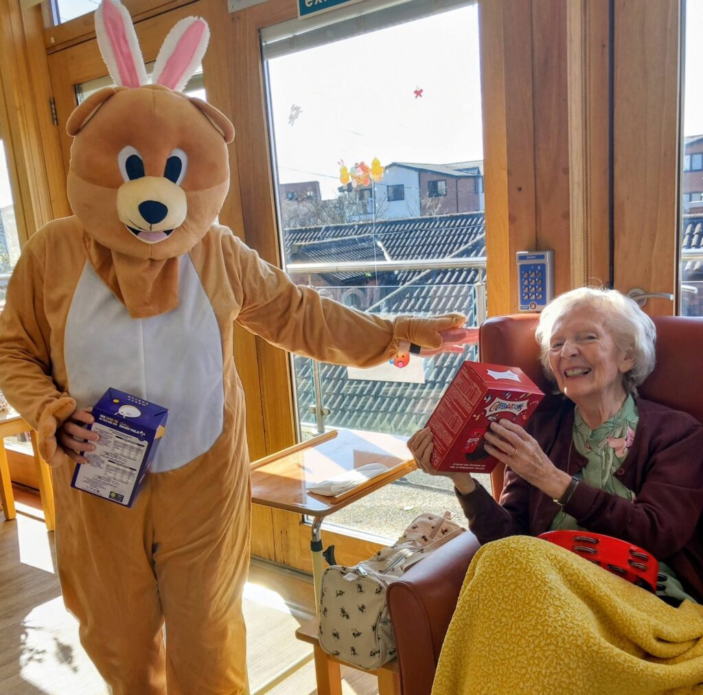 During an Easter celebration at Lear House, a person in a bunny costume hands a box of chocolates to an elderly woman seated in a chair beside a sunny window.