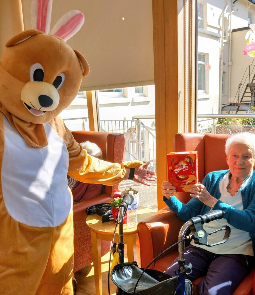 During an Easter celebration at Lear House, a person in a bunny costume stands beside an elderly woman sitting in a chair, who is holding a box of Maltesers.