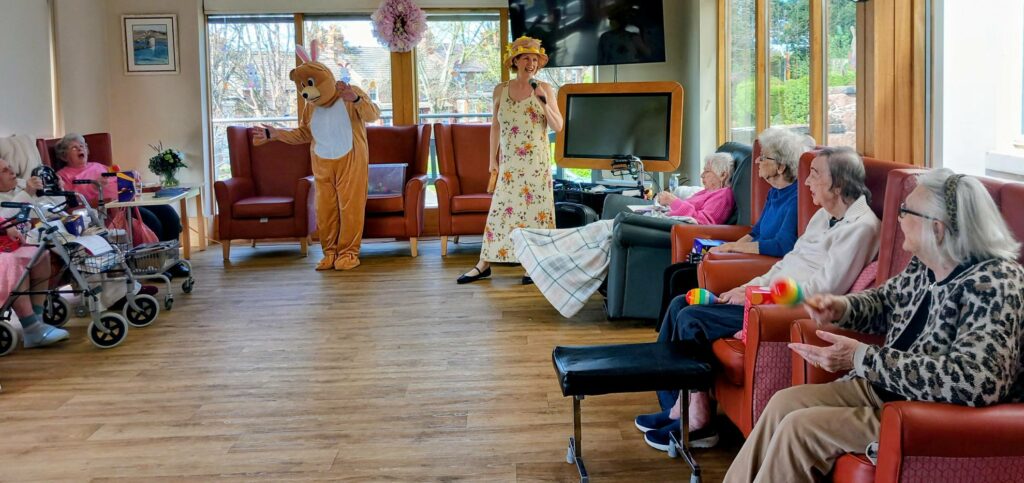 A person in a bear costume and another in a floral dress entertain elderly residents seated in armchairs during an Egg-stra Special Easter Celebration in Lear House’s brightly lit communal lounge.