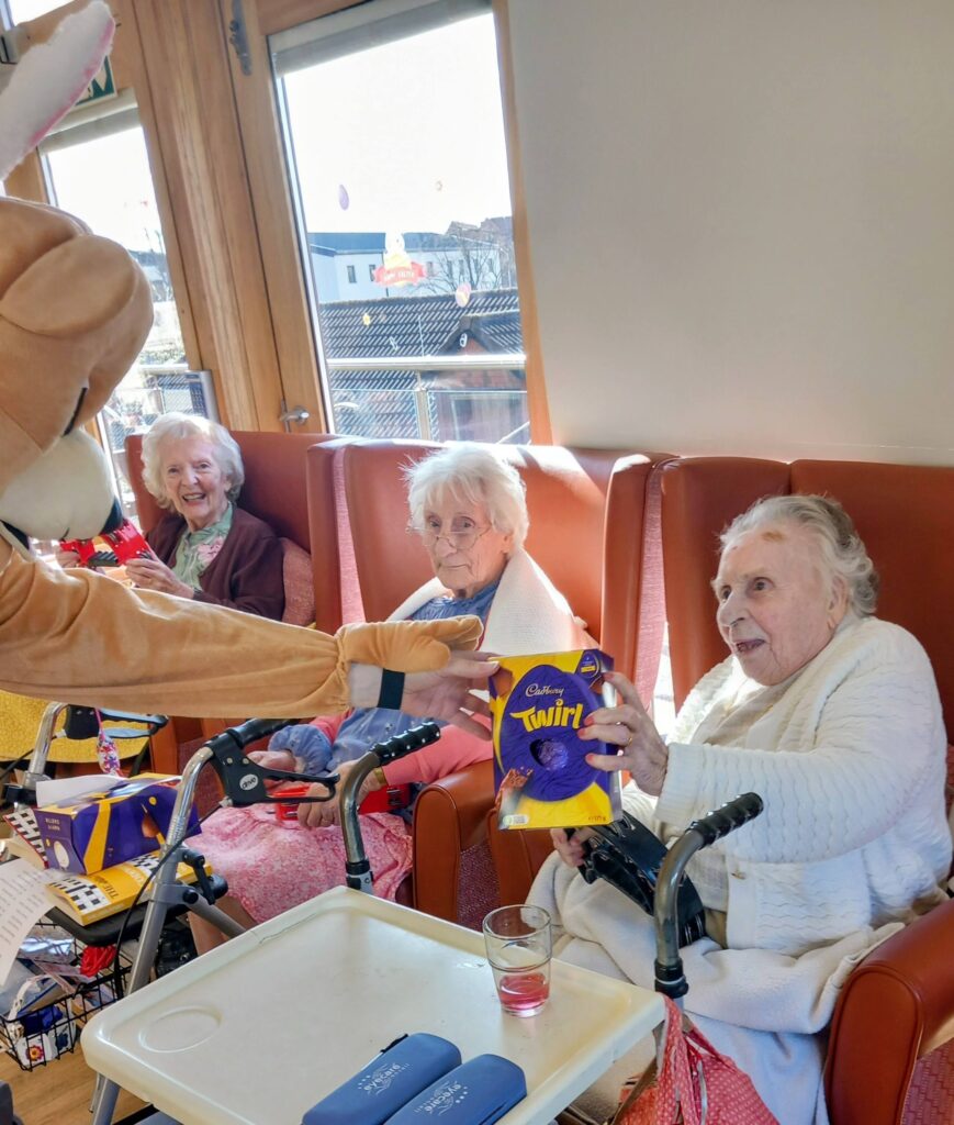 During an Egg-stra special Easter celebration at Lear House, three elderly women seated in chairs are given Cadbury Twirl Easter eggs by someone in a bunny costume inside a brightly lit room.