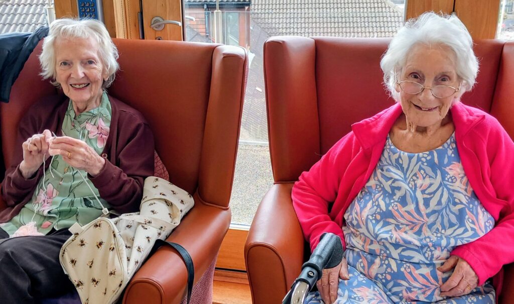 Two elderly women sit in armchairs by a window, smiling. One, holding some sewing, wears a floral blouse with a beige handbag; the other wears glasses, a pink jacket and a blue dress with a distinctive floral pattern.