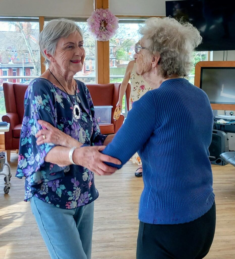 Two elderly women are holding hands and dancing together in a bright room at Lear House, enjoying an Egg-stra Special moment with chairs and a telly visible in the background.