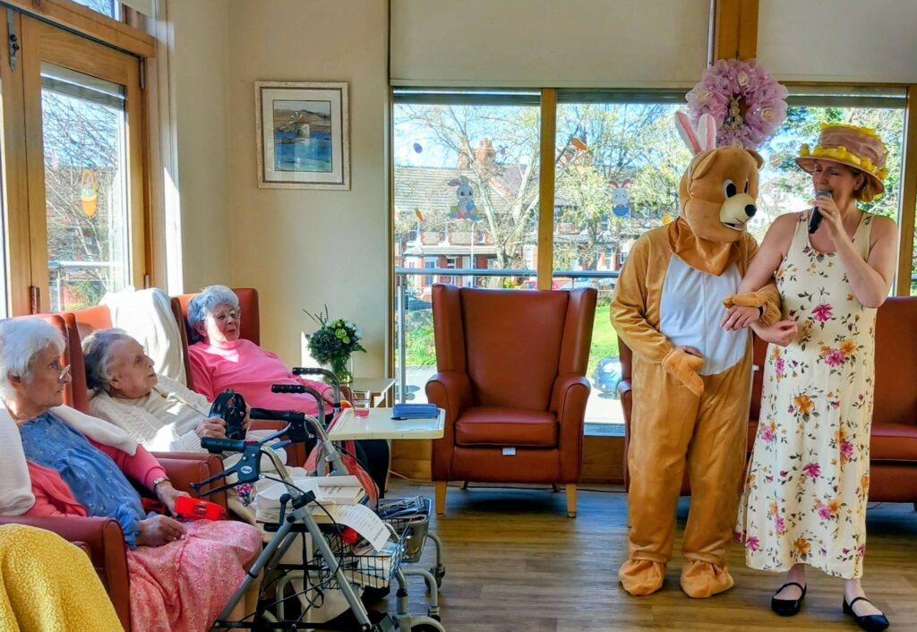 During an Easter celebration at Lear House, a group of elderly women relax in the lounge whilst someone dressed as the Easter Bunny and a woman in a floral dress and sunhat sing or speak into a microphone.