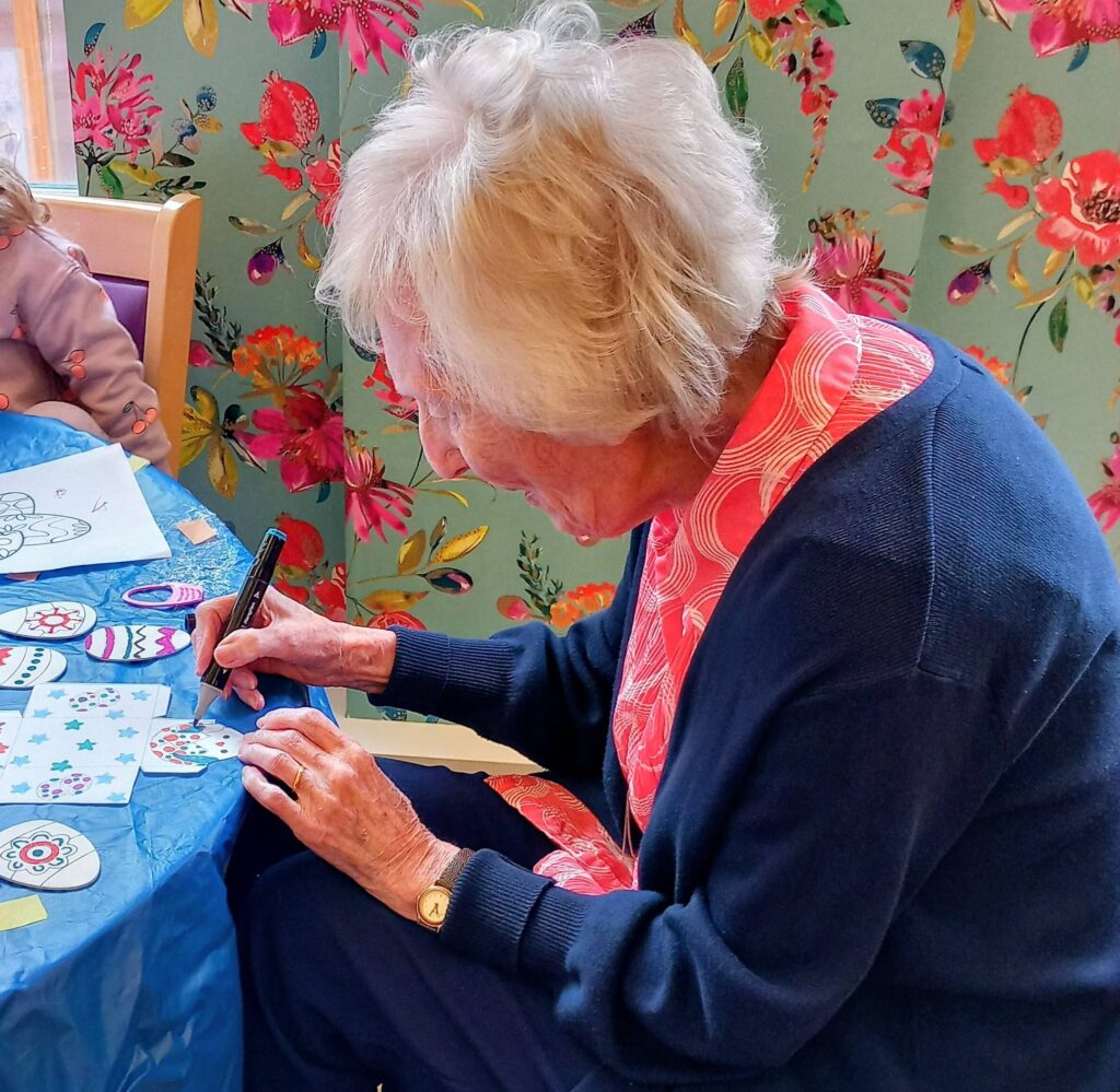 An elderly woman sits at a table, decorating paper crafts with a black marker. A child is partially visible next to her. The background features floral wallpaper.
