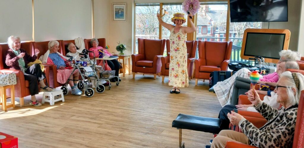 At Lear House, a group of elderly people sit in a lounge watching a person in a floral dress and hat perform with maracas during an Egg-stra Special Easter Celebration.