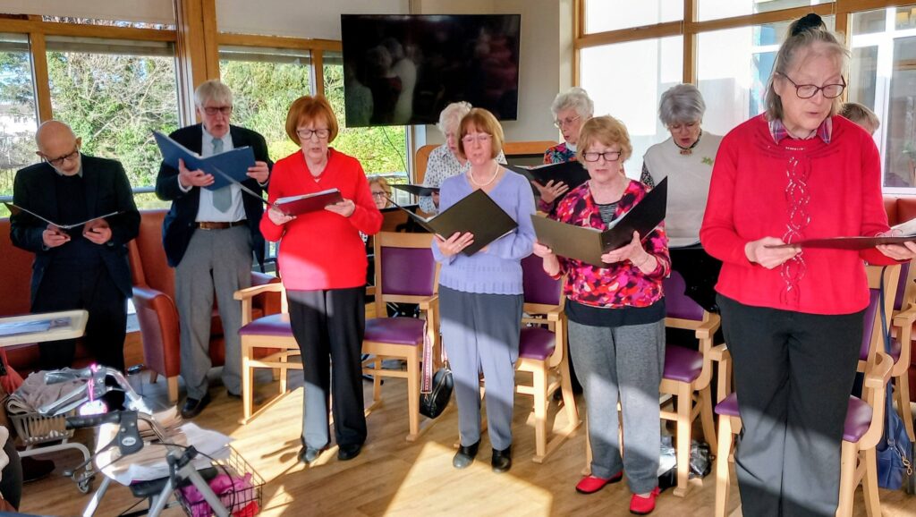 A group of elderly people stand in a sunlit room, holding songbooks and singing together, with chairs and large windows in the background.