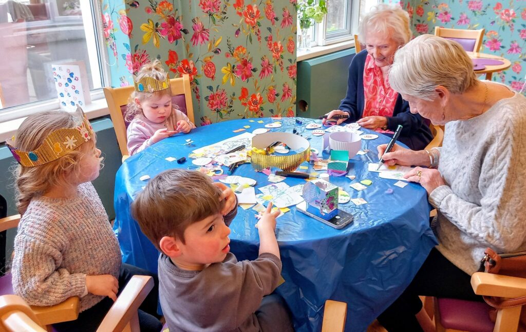 Two elderly women and three young children sit round a table covered with art materials, creating crafts together in a brightly decorated room.