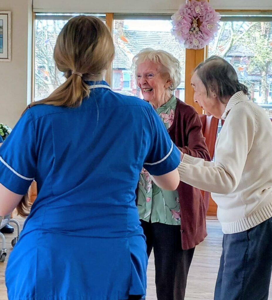 A carer in blue uniform assists two elderly women, smiling and holding hands, during an Egg-stra Special Easter Celebration at Lear House in a brightly lit room with large windows.