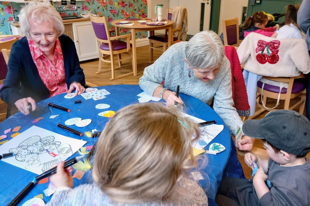 Two elderly women and two young children sit at a table covered with a blue cloth, colouring and drawing together with felt-tip pens and paper in a bright room.