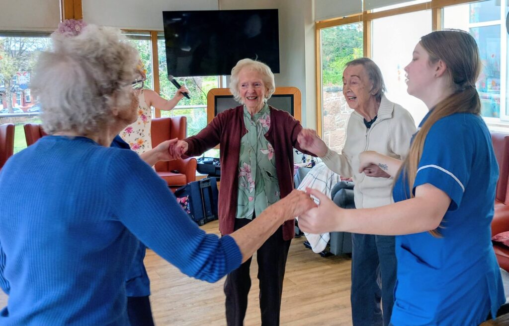 Four elderly women and a carer in uniform hold hands and dance in a brightly lit room at Lear House, enjoying an egg-stra special Easter celebration by the large windows, with a television on the wall.