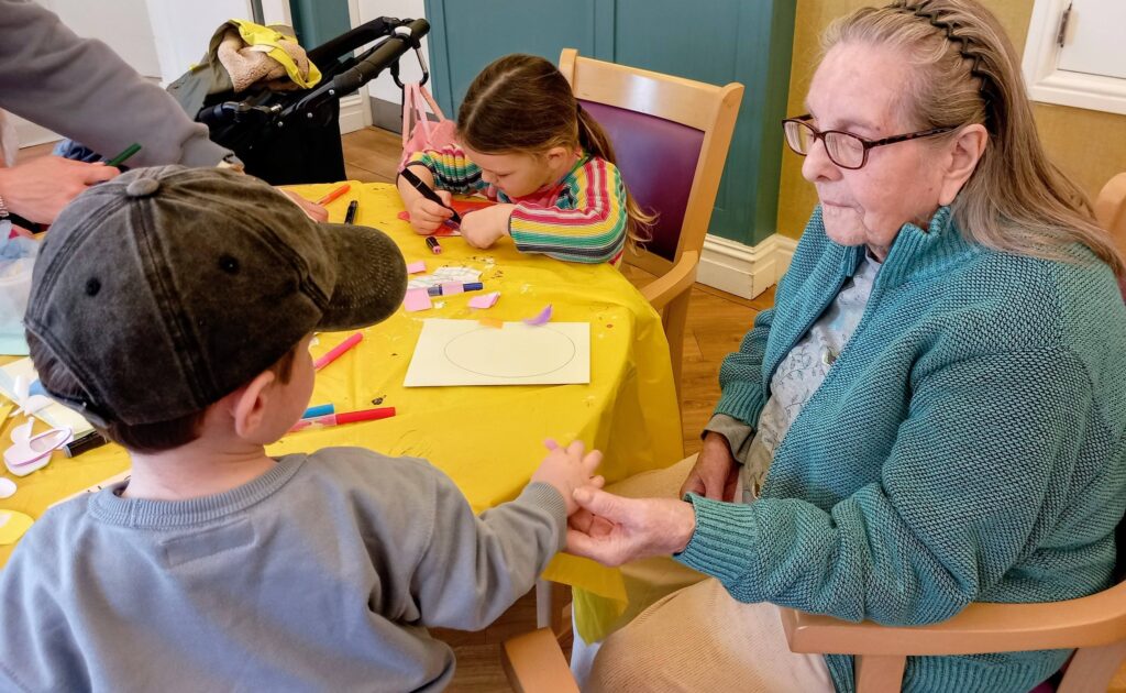 An elderly woman sits at a table holding a young boy's hand, whilst a girl works on a craft project using paper and felt-tip pens.