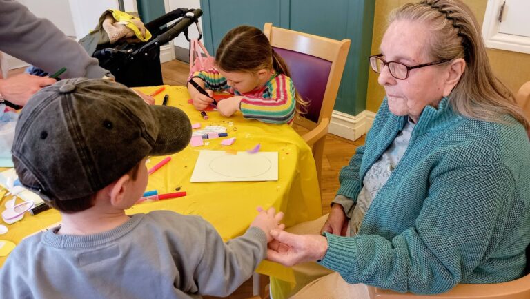 An elderly woman sits at a table holding a young boy's hand, whilst a girl works on a craft project using paper and felt-tip pens.