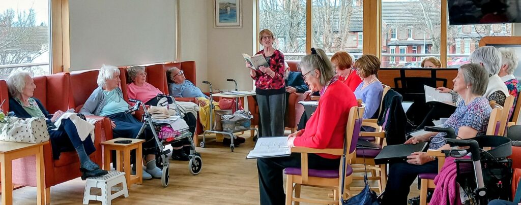A group of elderly people sit in a bright communal room, listening to a woman who is standing and reading aloud from a book.