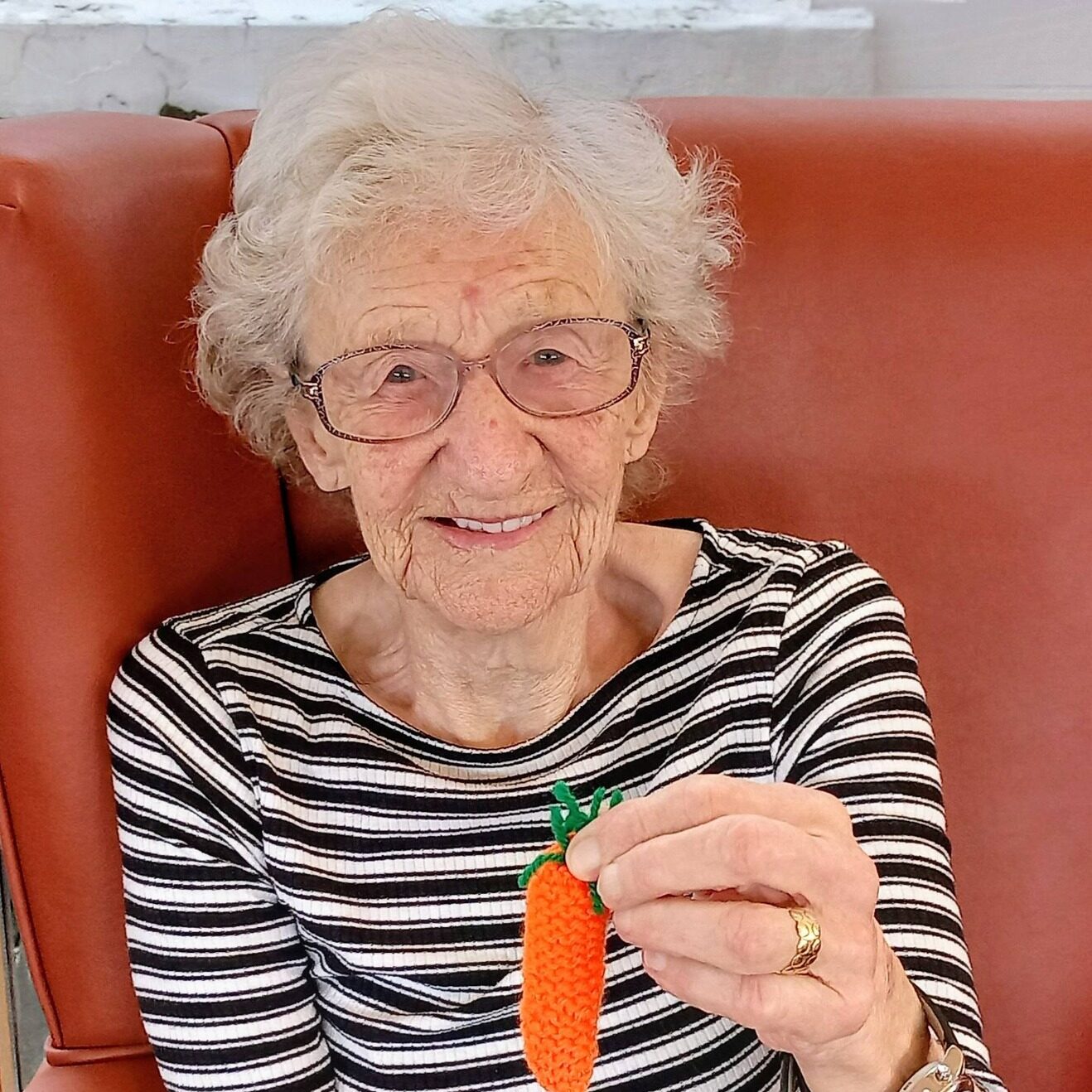 An elderly woman wearing glasses and a striped top sits in an orange chair, smiling and holding up a special crocheted carrot—her stitching skill makes it just right for Easter.