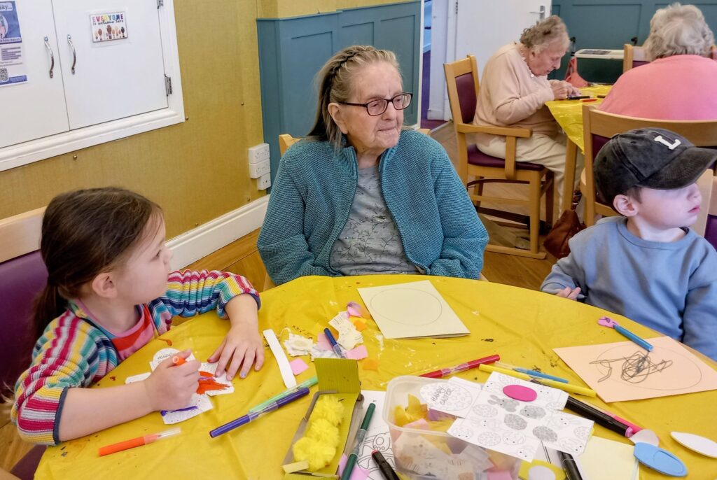 An elderly woman sits at a table with two young children, surrounded by art materials and drawings in a communal room.