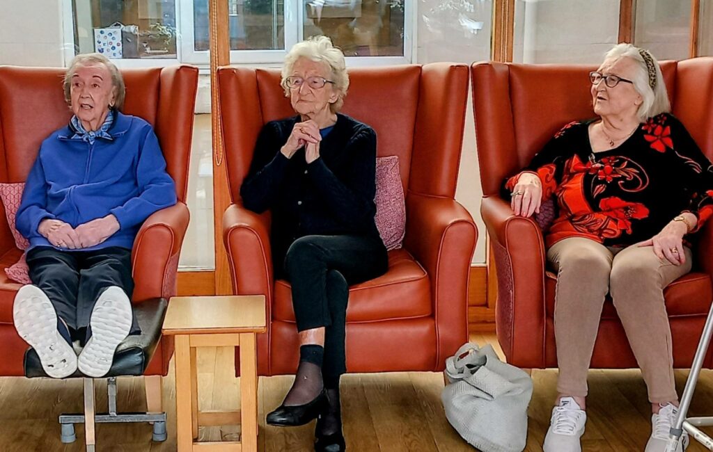 Three elderly women sit indoors in red armchairs, facing forwards. One is dressed in blue, one in black, and the other in black and red. A white bag rests on the floor beside them.