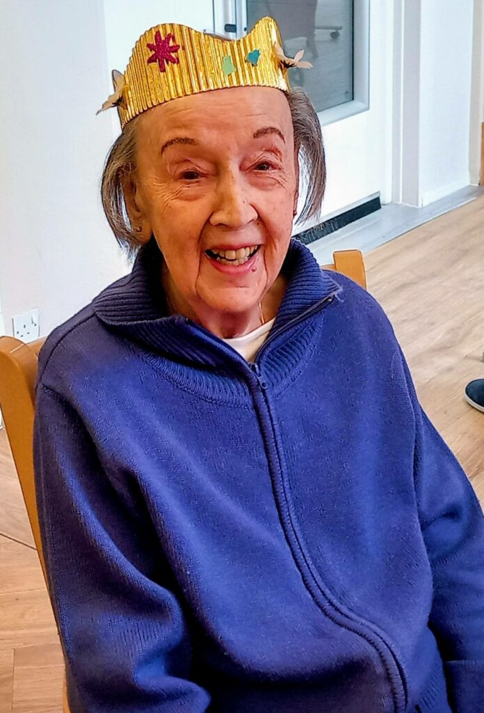 Elderly woman in a handmade yellow paper crown and a purple zip-up jumper, seated indoors and smiling at the camera.